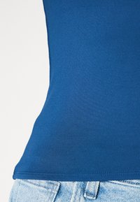 Μπλε εφαρμοστό αμάνικο μπλουζάκι από ελαστικό ύφασμα, με απλή υφή και φαρδιά λαιμού, συνδυασμένο με τζιν παντελόνι σε ανοιχτό χρώμα.