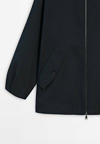 Manche de veste noire et bas avant avec fermeture éclair et poche à rabat inclinée sécurisée par un bouton sur fond blanc.