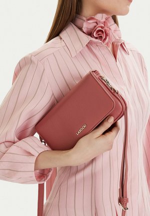 Frau in rosa gestreiftem Hemd mit Rosendetail, die eine strukturierte pinkfarbene Lasocki-Clutch mit Handgelenkriemen hält.