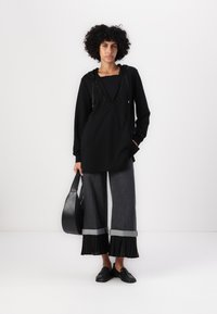 Felpa nera con collo decorato, abbinata a pantaloni grigi scuri a gamba larga con dettagli plissettati e orli arrotolati. La modella tiene una borsa nera.