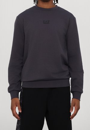 Personne portant un sweat-shirt à manches longues gris foncé avec le logo EA7 Emporio Armani sur la poitrine et un pantalon noir avec un détail de texte sur le côté.