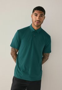 Teal polo skjorte med korte ærmer, to-knaps lukning og en lille logo detalje på brystet; iført med sorte bukser. Glat tekstur.