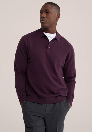 Homme portant un polo violet à manches longues par-dessus un sous-maillot blanc et un pantalon gris foncé, debout les mains dans les poches, regardant sur le côté.