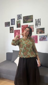 Γυναίκα με φλοράλ δαντελένιο τοπ και μαύρη φούστα αγγίζει τα μαλλιά της, στέκεται μπροστά σε γκρι καναπέ με κολάζ τέχνης και φωτογραφιών σε λευκό τοίχο.