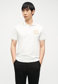 Άνδρας που φοράει λευκό πόλο με χρυσή λεπτομέρεια και λογότυπο, μαύρο παντελόνι, στέκεται με τα χέρια στις τσέπες, κοιτάζοντας προς τα πλάγια σε απλό φόντο.