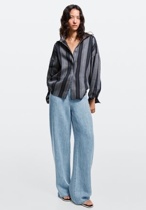 Woman wearing a loose dark striped button-up shirt with wide light blue denim pants and black sandals, standing against a plain background.