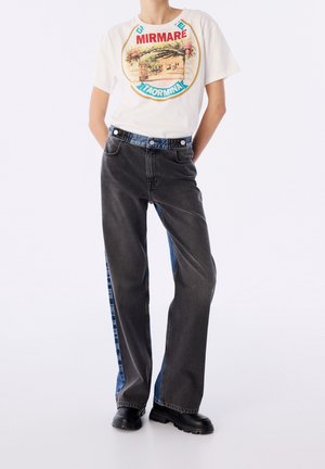 Person wearing white graphic tee with "MIRMARE TAORMINA" text and black jeans with blue side stripes, standing with hands behind back.