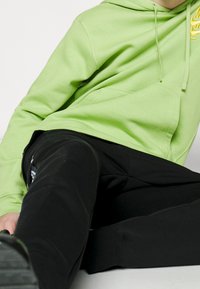 Limegrön huvtröja med dragsnöre och en känguruficka, kombinerad med svarta sweatpants med en sidologga. Slät, avslappnad tyg.