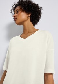 Top s V výstřihem a krátkým rukávem v barvě off-white, s volným střihem a hladkou texturou, se zdobením na bočních švech a minimalisticým designem.