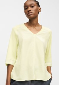 Νέα γυναίκα με πλεκτά μαλλιά που φοράει ανοιχτοκίτρινη μπλούζα με V-λαιμόκοψη και μανίκια τριών τετάρτων, χέρια στις τσέπες, ουδέτερο φόντο.