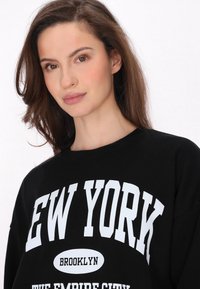 Fekete pulóver, amelyet merész fehér betűk díszítenek: "NEW YORK BROOKLYN A BIRODALOM VÁROSA." Kerek nyakú és puha, sima tapintású.