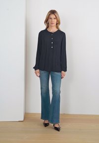 Blouse noire à manches longues avec un col rond et une patte de boutonnage, associée à un jean bleu évasé et des chaussures à talons noires. Design simple, texture lisse.