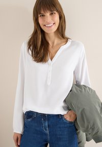 Blouse blanche à manches longues avec un col en V et une patte de boutonnage, associée à un jean bleu. Une veste verte claire est posée sur un bras.