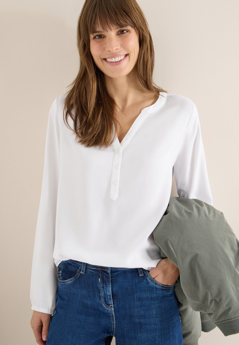 Blouse blanche à manches longues avec un col en V et une patte de boutonnage, associée à un jean bleu. Une veste verte claire est posée sur un bras.