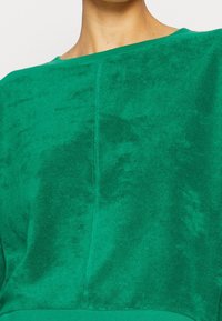 Zelený pulovr s jemnou, sametovou texturou. Má široký výstřih a surové švy, které zdůrazňují jeho uvolněný, moderní design.