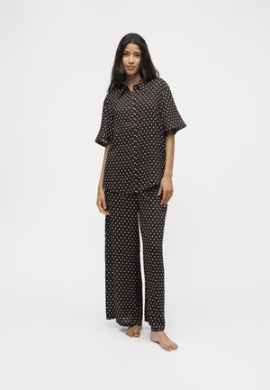 Femme debout pieds nus portant une chemise noire à manches courtes boutonnée et un pantalon large à pois blancs sur un fond uni.