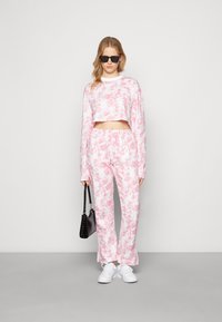 Sweatshirt cropped em tie-dye rosa e branco e calças de fato combinando, acompanhados de ténis brancos e uma mala preta. Textura de tecido suave.