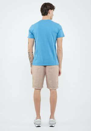 Uomo con braccio tatuato che indossa una maglietta blu, pantaloncini beige al ginocchio e scarpe da ginnastica bianche, in piedi di spalle su uno sfondo semplice.