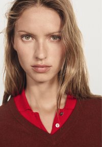 Maglione marrone a scollo a v sopra una camicia rossa con colletto. La texture sembra morbida; la camicia ha due bottoni. Il modello ha lunghi capelli mossi e un'espressione neutra.