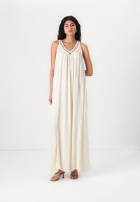 Robe maxi en tissu ivoire clair, présentant un décolleté en V avec des détails en crochet. Design fluide avec une texture douce, associée à des sandales à talons.