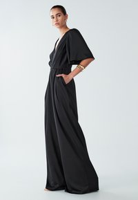 Sieviete valkā melnu plašu kombinezonu ar dziļu V veida izgriezumu un īsām plīvuru piedurknēm, papildināta ar zelta rotaslietām un auskariem ar gredzeniem.