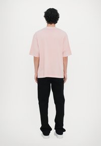T-shirt de algodão rosa com mangas curtas, decote redondo e ajuste descontraído, combinada com calças pretas e ténis brancos, vista de trás.