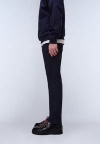 Mand stående sidelæns iført sorte bukser, sorte kraftige sko med røde snørebånd, mørkeblå trøje og lysegrå undertrøje på hvid baggrund.