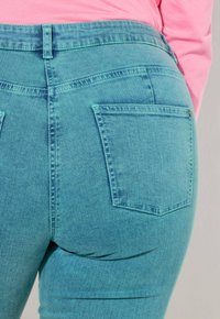 Jeansy z denimu w kolorze morskim z wysokim pasem, wyposażone w dwie tylne kieszenie i kontrastujące szwy. Tkanina wydaje się miękka z delikatną fakturą.