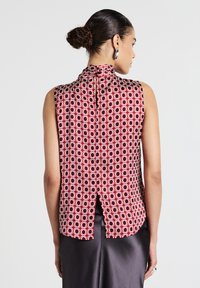 Donna con i capelli raccolti a chignon, che indossa una blusa rossa senza maniche con motivo e uno spacco sul retro, insieme a una gonna di satin scuro, in piedi su uno sfondo neutro.