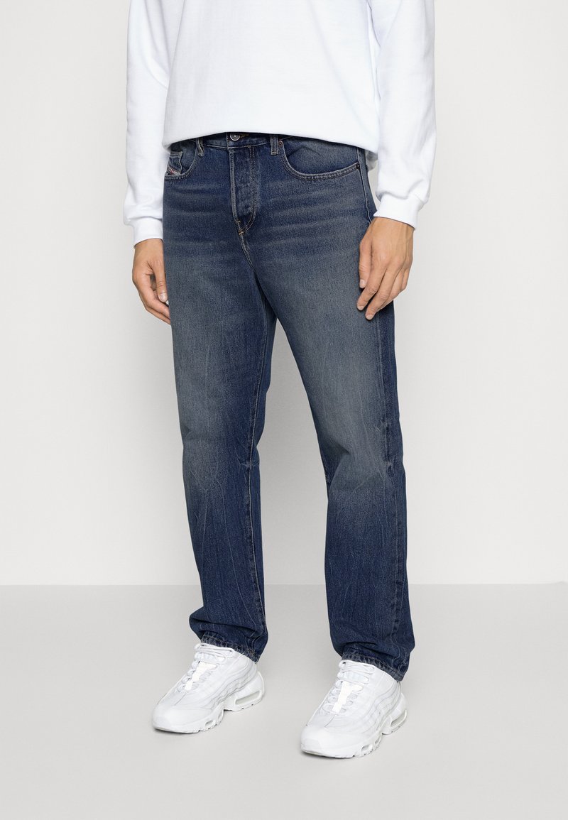 Dunkelblaue Jeans aus Denim mit einer lockeren Passform, fünf Taschen und sichtbaren Stickdetails. Getragen mit einem weißen Langarmshirt und weißen Sneakers.