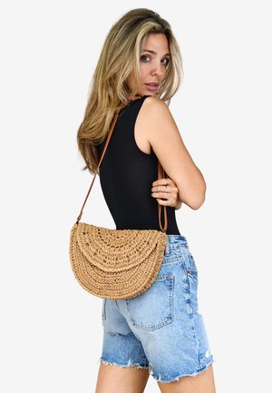 Bolso de paja tejido tipo bandolera con una silueta curvada, correa de cuero marrón y detalles en costura decorativa, combinado con pantalones cortos de mezclilla.