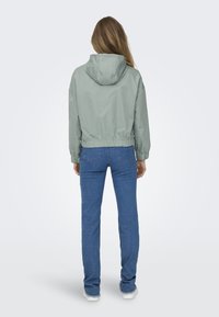 Lys grøn cropped jakke med hætte, elastisk talje og lange ærmer, parret med blå jeans og hvide sko. Enkel design og glat tekstur.