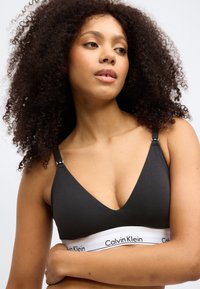 Crna pamučna braleta s dubokim V-izrezom i podesivim trakama. Ima bijelu elastičnu traku s "Calvin Klein" logotipom na dnu.