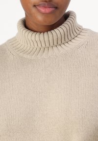 Beige Rollkragenpullover mit geripptem Kragen und strukturiertem Strickmuster, aus einem weichen, glatten Material. Nahaufnahme von Ausschnitt und Brust.