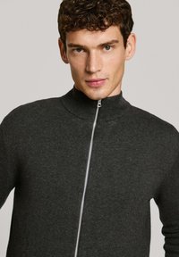 Grå stickad zip-up-tröja med hög krage och silverdragkedja. Har en slät textur och en åtsittande design. Nära bild av överkroppen.