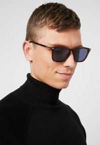 Sköldpaddsformade solglasögon med mörka linser, metallaccentuerade, rektangulär form. Modellen bär en svart polotröja. Neutral bakgrund.