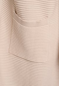 Tela beige acanalada con un bolsillo frontal. La textura es suave y ligeramente elástica, con finas líneas horizontales. Diseño sencillo y funcional.