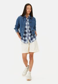 Jeansowa kurtka z zapięciem na guziki i dwiema kieszeniami, noszona na niebieskiej koszuli w kratę; w połączeniu z jasnymi szortami i lekkimi trampkami. Białe tło.