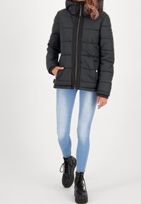 Persoon in een zwarte gewatteerde jas, lichtblauwe skinny jeans en zwarte veterschoenen op een platform, staand met één hand in een zak.