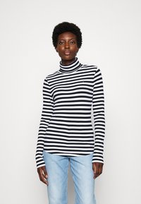 Black and white striped turtleneck top with long sleeves, ribbed texture, and a small logo on the chest, paired with light blue jeans.