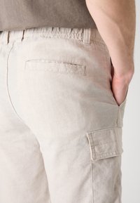 Lys beige cargo bukser laget av et mykt stoff, med en baklomme og elastisk midje. Designet inkluderer en sidelomme til cargo.