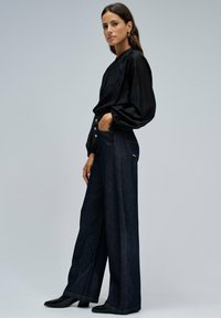 Donna in piedi con una mano in tasca, indossa una blusa nera con maniche a palloncino, jeans a gamba larga scuri a vita alta e scarpe con tacco nere su sfondo grigio.