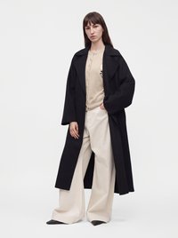 Femme portant un long manteau noir par-dessus un pull beige boutonné et un pantalon large crème, debout avec une main dans la poche devant un fond uni.