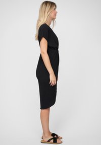 Black knee-length dress with short sleeves, soft texture, and a knotted detail at the waist. Paired with black flat sandals.