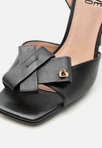 Μαύρο δερμάτινο πέδιλο με ανοιχτά δάχτυλα και τακούνι, με σταυρωτά λουριά και μικρό χρυσό διακοσμητικό σε σχήμα καρδιάς σε ένα λουρί.