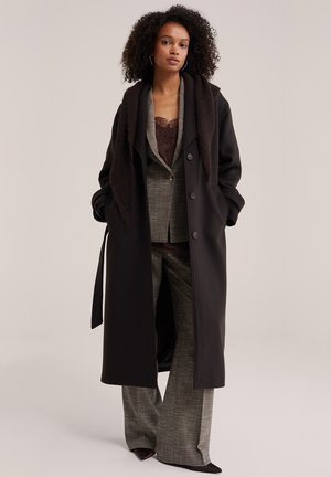 Femme aux cheveux bouclés portant un long manteau sombre par-dessus un costume à carreaux gris et un haut en dentelle marron, debout les mains dans les poches du manteau.