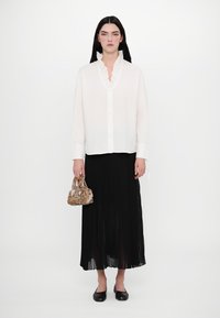 Weiße Bluse mit langen Ärmeln und Rüschenausschnitt, kombiniert mit einem schwarzen, gefalteten Maxirock. Ergänzt durch eine gemusterte kleine Handtasche und schwarze Ballerinas.