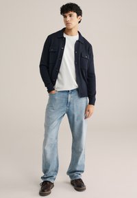 Marinblå stickad cardigan med knappstängning, två bröstfickor, bärs över en vit t-shirt, kombinerad med ljusblå jeans och mörkbruna skor.