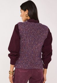 Gilet lavorato a maglia viola con sfumature multicolori, caratterizzato da una texture a coste e un collo alto, indossato sopra una camicetta bordeaux con maniche a sbuffo.