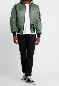 Uomo che indossa una giacca bomber verde, una t-shirt bianca, pantaloni neri e sneakers alte nere, in piedi davanti a uno sfondo bianco uniforme.